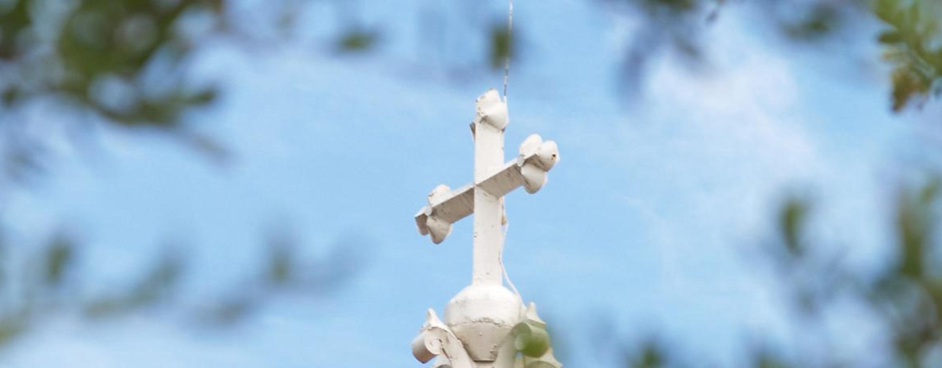 The image depicts a white cross atop a building, set against a blue sky. The cross has decorative elements and is viewed through a soft focus of green leaves, creating a frame around it. The scene conveys a peaceful and serene atmosphere, highlighting the cross as a central symbol of faith and spirituality.